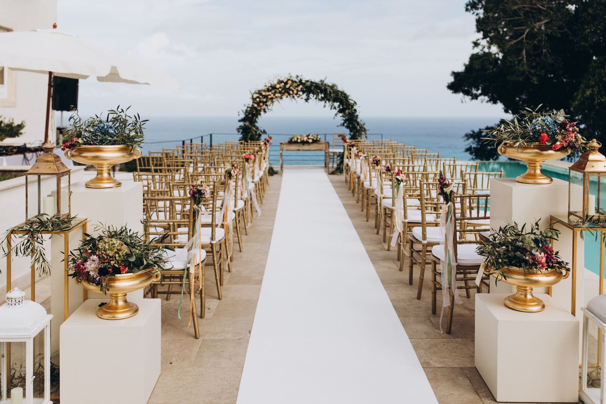 A seaside ceremony aisle lined with chairs and gold flower stands, leading to a floral arch by the ocean