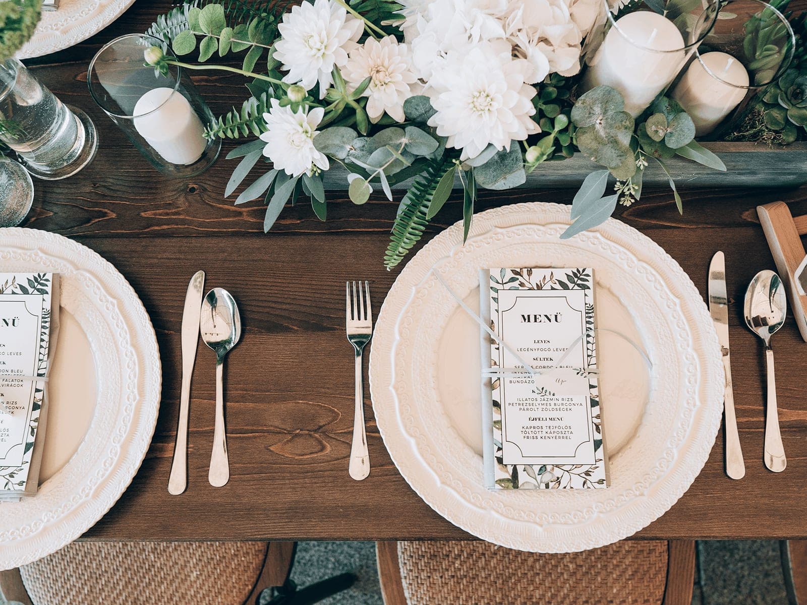 A reception table set with white plates, menus and a centrepiece of white flowers and greenery
