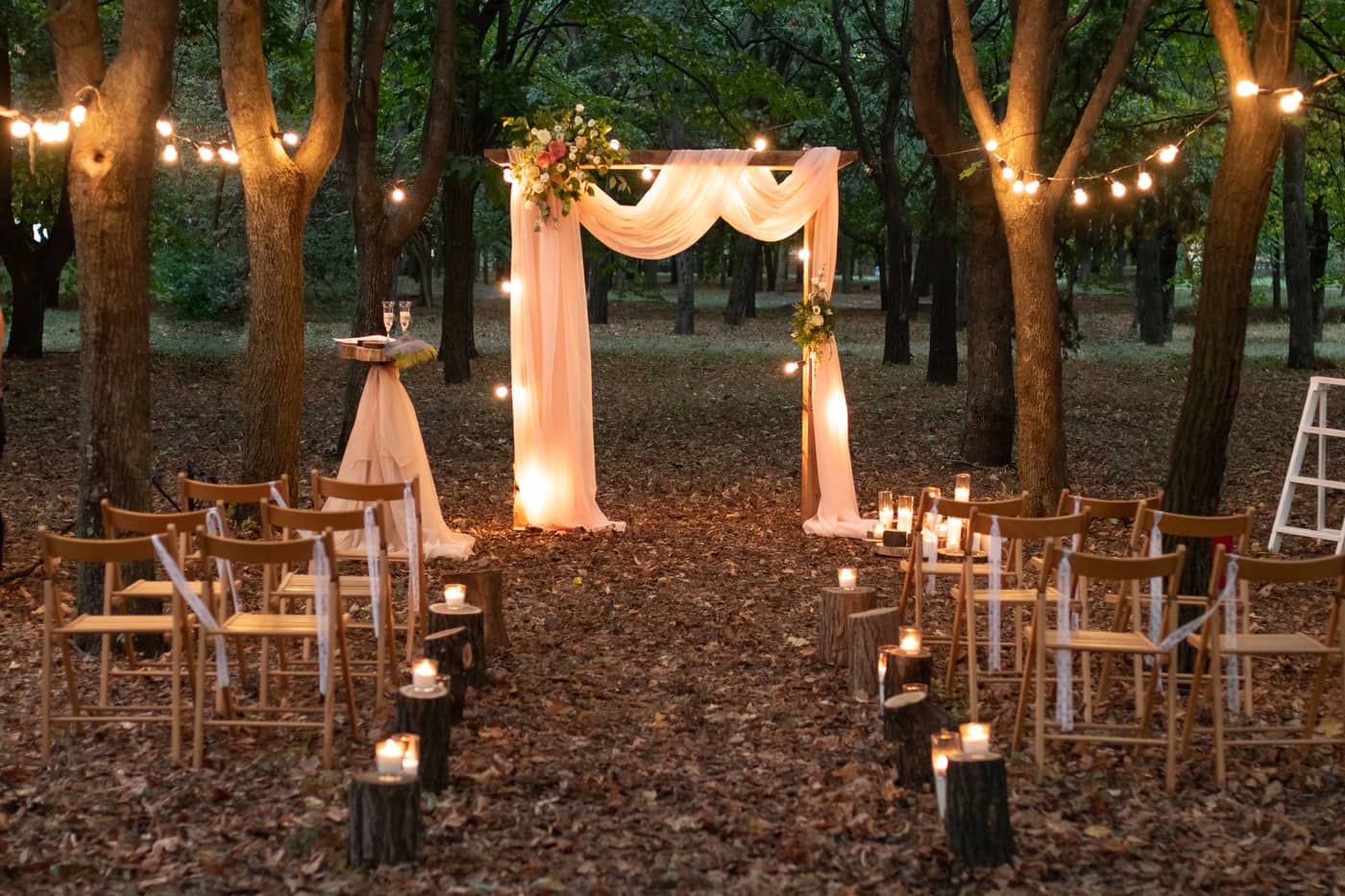 An outdoor forest ceremony with draped fabric arch, string lights, candles and wooden chairs