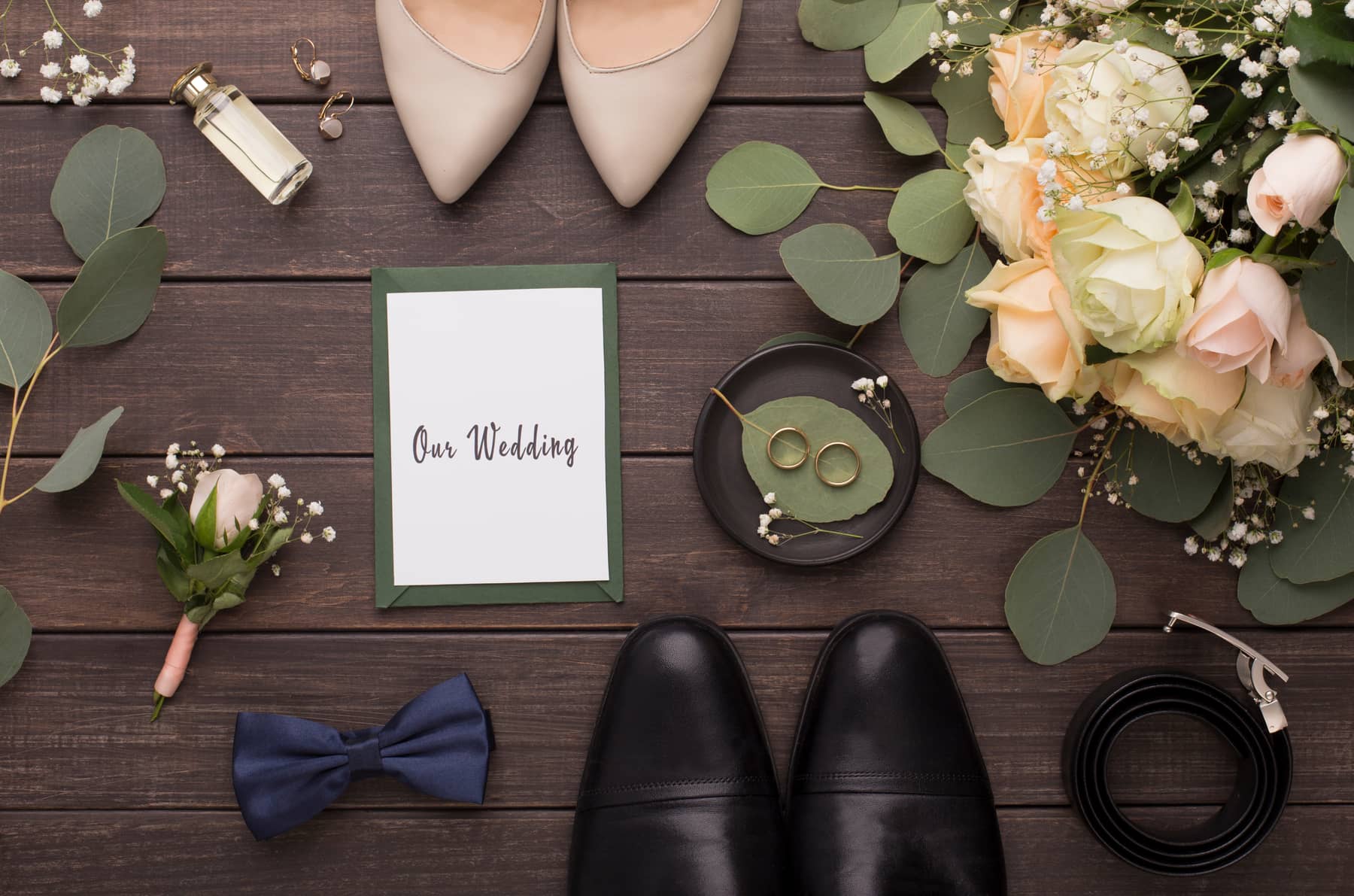 A flat lay of wedding shoes, a bow tie, rings and a card reading “Our Wedding” beside cream roses